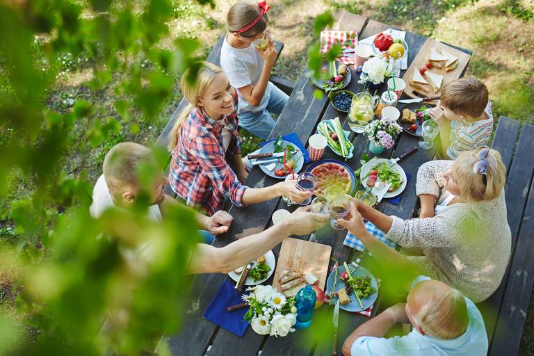 Perheillallinen ulkona, pöydän ympärillä aikuisia kerääntyneenä nauttimaan ruokaa ja juomaa.